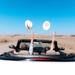 Two people in a convertible automobile on the road throwing up their hats